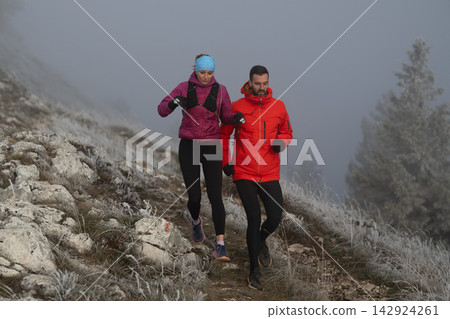 Couple Trail Running Together On A Foggy Mountain Path In Winter Jackets Couple Trail Running Together On A Foggy Mountain Path In Winter Jackets 142924261