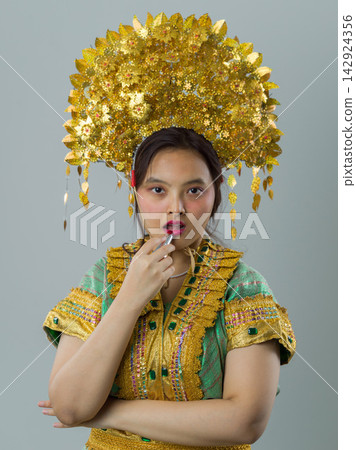 Young Indonesian Asian Woman Wearing Ornate Gold Headdress and Traditional Ceremonial Costume Applying Lipstick Young Indonesian Asian Woman Wearing Ornate Gold Headdress and Traditional Ceremonial Costume Applying Lipstick 142924356