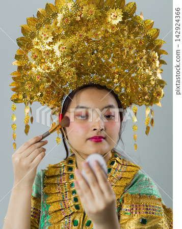 Indonesian Asian Girl Applying Makeup Wearing Ornate Traditional Golden Headdress and Embellished Costume Indonesian Asian Girl Applying Makeup Wearing Ornate Traditional Golden Headdress and Embellished Costume 142924366