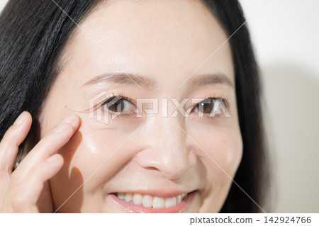 一位中年日本女性面帶微笑地檢查著眼周的皺紋;這是一張針對 50 多歲女性的抗衰老護理和護膚圖片。 一位中年日本女性面帶微笑地檢查著眼周的皺紋;這是一張針對 50 多歲女性的抗衰老護理和護膚圖片。 142924766