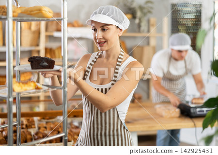 Young saleswoman putting pastry on rack in bakery Young saleswoman putting pastry on rack in bakery 142925431