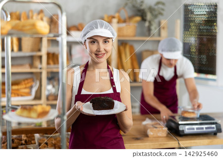 Smiling saleswoman presenting slice of chocolate cake in bakery Smiling saleswoman presenting slice of chocolate cake in bakery 142925460