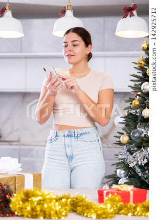 Young European woman in casual clothes surfing Internet on phone next to wrapped gift and Christmas tree before holidays in living room Young European woman in casual clothes surfing Internet on phone next to wrapped gift and Christmas tree before holidays in living room 142925712