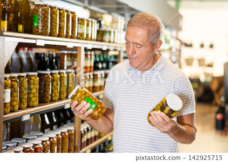 Elderly man choosing pickled olives in jars in grocery store Elderly man choosing pickled olives in jars in grocery store 142925715