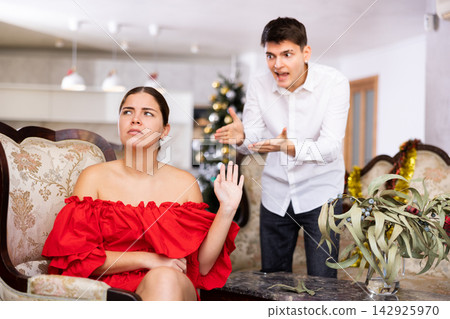 Emotional distressed young male and female screaming at each other while sitting on couch in front of Christmas tree in apartment 142925970