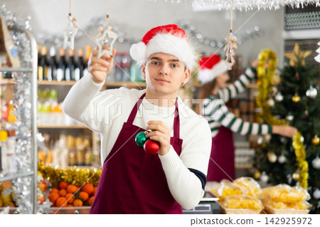 Young guy seller decorates grocery store for christmas Young guy seller decorates grocery store for christmas 142925972