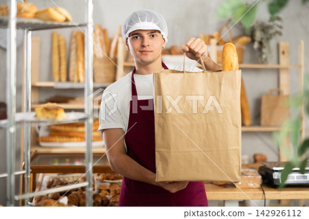 Young bakery clerk holding paper bag with fresh baked goods Young bakery clerk holding paper bag with fresh baked goods 142926112