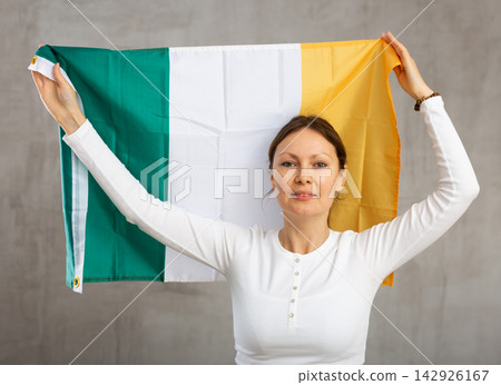 Happy young woman holding flag of Ireland against unicoloured background Happy young woman holding flag of Ireland against unicoloured background 142926167