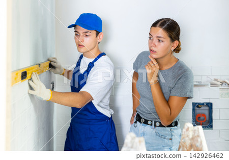 Young contractor with female homeowner checking wall flatness with level meter Young contractor with female homeowner checking wall flatness with level meter 142926462