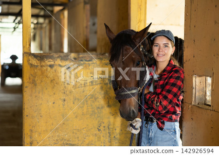 Smiling young girl in plaid shirt and gloves taking care and talking to horse on ranch Smiling young girl in plaid shirt and gloves taking care and talking to horse on ranch 142926594