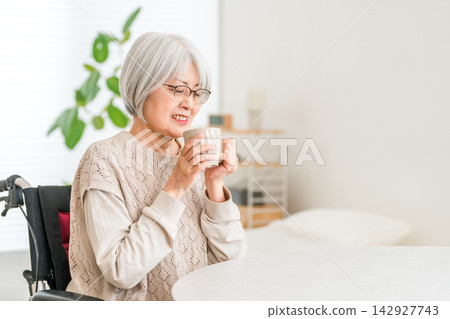 一位坐在輪椅上的老婦人在家中一邊喝著日本茶一邊放鬆。 一位坐在輪椅上的老婦人在家中一邊喝著日本茶一邊放鬆。 142927743