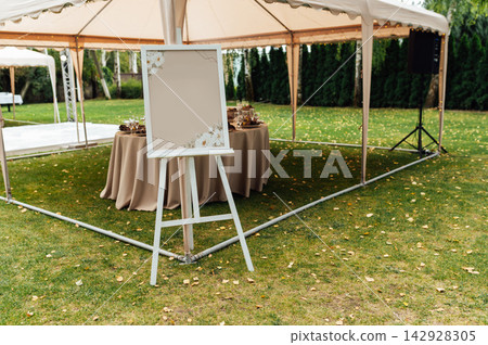 Blank wedding welcome sign board mockup on white easel under marquee tent outdoors. Blank wedding welcome sign board mockup on white easel under marquee tent outdoors. 142928305