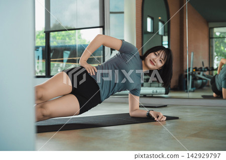 Young woman wearing workout clothes doing side plank exercise on yoga mat indoors in modern gym with natural light Young woman wearing workout clothes doing side plank exercise on yoga mat indoors in modern gym with natural light 142929797