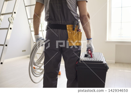 Rear view of electrician with toolbox and cable in bright renovation room Rear view of electrician with toolbox and cable in bright renovation room 142934650