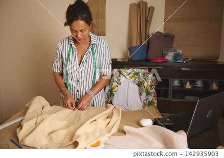 Woman seamstress tailoring a shirt in a home studio with measuring tape and laptop Woman seamstress tailoring a shirt in a home studio with measuring tape and laptop 142935903