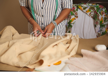 Woman tailor measuring and hand-sewing a beige shirt in a small tailor shop 142935904
