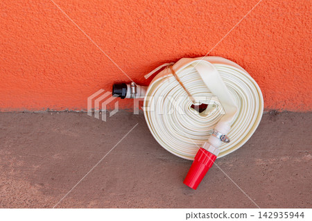 Rolled white hose with nozzle against orange wall. Firefighting equipment stored for emergency use. Industrial water supply tool for safety and fire protection in residential building. Rolled white hose with nozzle against orange wall. Firefighting equipment stored for emergency use. Industrial water supply tool for safety and fire protection in residential building. 142935944