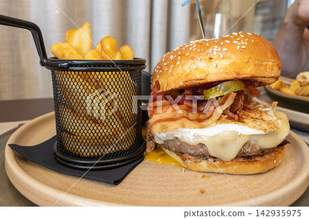 Gourmet hamburger with bacon, cheese, and pickles served with crispy fries in a restaurant setting Gourmet hamburger with bacon, cheese, and pickles served with crispy fries in a restaurant setting 142935975