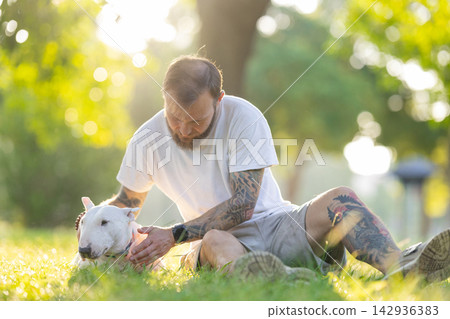 Old bull terrier sits on grass while male owner with tattoos gently pets the dog in a sunlit park setting surrounded by trees and soft greenery Old bull terrier sits on grass while male owner with tattoos gently pets the dog in a sunlit park setting surrounded by trees and soft greenery 142936383