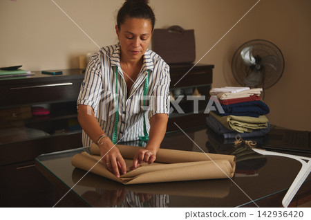 Woman folding fabric and preparing pattern in a small tailoring studio for a clothing business Woman folding fabric and preparing pattern in a small tailoring studio for a clothing business 142936420