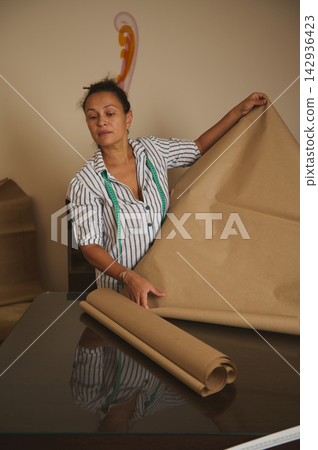 Woman unrolling kraft paper on table with measuring tape preparing patterns for sewing and crafting Woman unrolling kraft paper on table with measuring tape preparing patterns for sewing and crafting 142936423