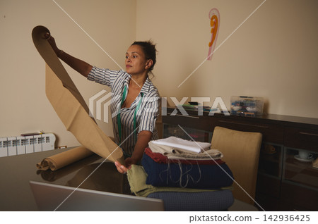 Woman preparing pattern paper and fabrics for sewing in a home studio, focused small business tailor Woman preparing pattern paper and fabrics for sewing in a home studio, focused small business tailor 142936425