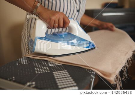 Woman ironing a light scarf on an ironing board at home with a steam iron Woman ironing a light scarf on an ironing board at home with a steam iron 142936458
