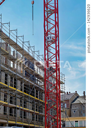 Red construction crane near concrete building under construction with industrial scaffolding against blue sky 142936620