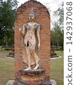 An elegant Sukhothai-style walking Buddha statue stands in front of a brick wall in Sukhothai Historical Park, Thailand. 142937068