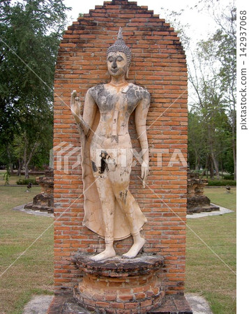 An elegant Sukhothai-style walking Buddha statue stands in front of a brick wall in Sukhothai Historical Park, Thailand. An elegant Sukhothai-style walking Buddha statue stands in front of a brick wall in Sukhothai Historical Park, Thailand. 142937068