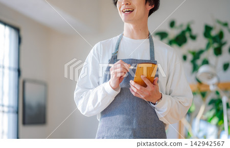 一名身穿圍裙的男子麵帶微笑地邊做筆記邊講話。 一名身穿圍裙的男子麵帶微笑地邊做筆記邊講話。 142942567