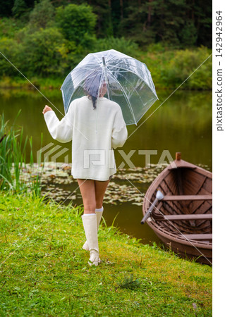 Young woman in white sweater and knee-high socks walks with a clear umbrella by a tranquil lake, wooden boat moored nearby, surrounded by lush greenery Young woman in white sweater and knee-high socks walks with a clear umbrella by a tranquil lake, wooden boat moored nearby, surrounded by lush greenery 142942964