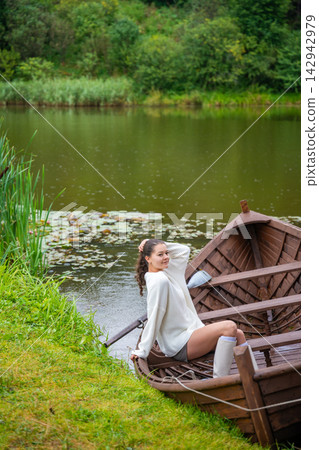 Young woman relaxing in wooden rowboat by lake on autumn day. Cozy sweater and rubber boots represent seasonal outdoor lifestyle and slow living in nature Young woman relaxing in wooden rowboat by lake on autumn day. Cozy sweater and rubber boots represent seasonal outdoor lifestyle and slow living in nature 142942979