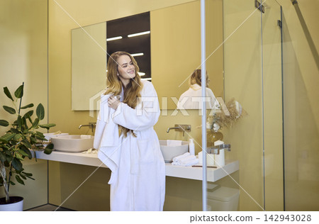 Smiling woman in bathrobe by bathroom mirror after shower, wellness routine Smiling woman in bathrobe by bathroom mirror after shower, wellness routine 142943028