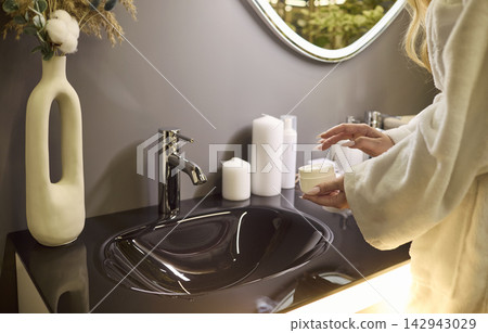 Woman in robe applying face cream at bathroom sink, wellness skincare routine Woman in robe applying face cream at bathroom sink, wellness skincare routine 142943029