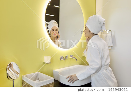 Woman in robe touching face at bathroom mirror, skincare and wellness daily Woman in robe touching face at bathroom mirror, skincare and wellness daily 142943031