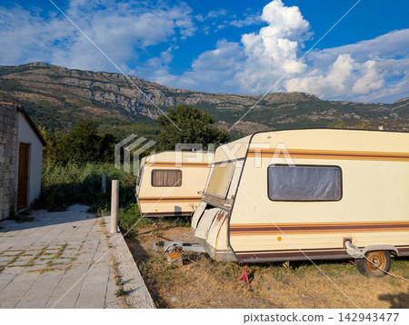 Two parked travel trailers beside small rural stone building on grassy roadside lot under blue sky with distant mountain scenery and scattered clouds with warm sunlight Two parked travel trailers beside small rural stone building on grassy roadside lot under blue sky with distant mountain scenery and scattered clouds with warm sunlight 142943477