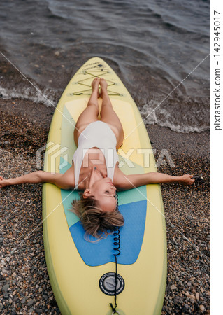 Relaxed woman lying on paddleboard by rocky shoreline wearing white swimsuit during outdoor leisure activity 142945017