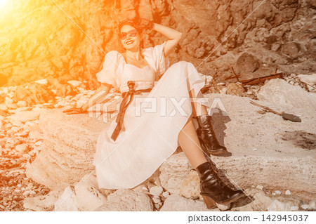 Woman beach fashion sits on rugged coastal rocks wearing a white vintage dress and boots during a golden sunset Woman beach fashion sits on rugged coastal rocks wearing a white vintage dress and boots during a golden sunset 142945037