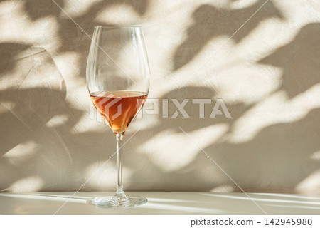 Elegant wine glass filled with rose beverage placed on white surface with dappled light. Elegant wine glass filled with rose beverage placed on white surface with dappled light. 142945980