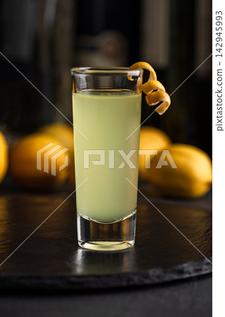 A tall shot glass filled with a pale green liquid and garnished with a lemon peel on a dark surface with lemons in the background A tall shot glass filled with a pale green liquid and garnished with a lemon peel on a dark surface with lemons in the background 142945993