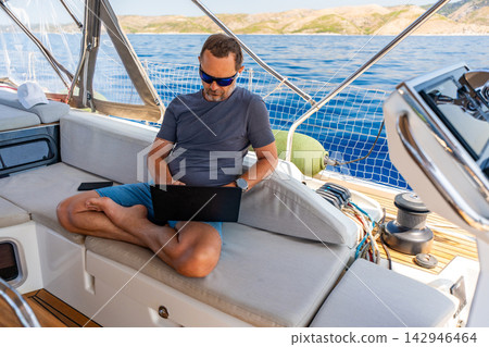 Focused man working on laptop aboard a sailing yacht. Productive remote office in a unique travel environment. 142946464