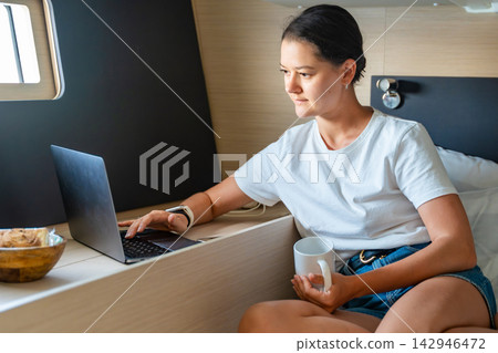 Young woman using a laptop and drinking coffee while working inside a comfortable sailing yacht cabin. Digital nomad lifestyle, remote work, freedom, travel and living aboard a sailboat. Young woman using a laptop and drinking coffee while working inside a comfortable sailing yacht cabin. Digital nomad lifestyle, remote work, freedom, travel and living aboard a sailboat. 142946472