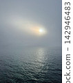 Glowing sun breaking through thick overcast fog over calm ocean water, creating a atmospheric moody seascape. 142946483