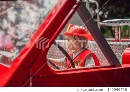 Little boy dressed as firefighter playing in a red toy fire truck at an outdoor playground with helmet safety vest pretend rescue game childhood dreams role play and looking at camera. Little boy dressed as firefighter playing in a red toy fire truck at an outdoor playground with helmet safety vest pretend rescue game childhood dreams role play and looking at camera. 142946726