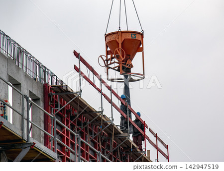 Nitra, Slovakia - February 25, 2025: Concrete hopper suspended above workers at a building construction site Nitra, Slovakia - February 25, 2025: Concrete hopper suspended above workers at a building construction site 142947519