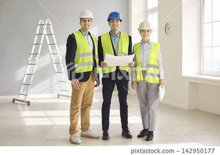 Confident engineers and contractors review blueprints in safety gear indoors Confident engineers and contractors review blueprints in safety gear indoors 142950177