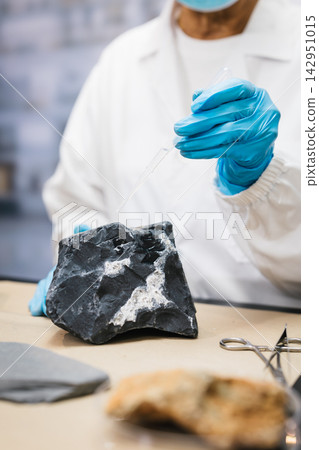 Scientist in a white lab coat and blue gloves uses a pipette to examine a dark rock sample on a laboratory table. Scientist in a white lab coat and blue gloves uses a pipette to examine a dark rock sample on a laboratory table. 142951015