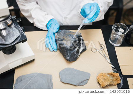 Geologist in a white lab coat and blue gloves examines a dark rock sample with a dropper beside a microscope and mineral specimens. Geologist in a white lab coat and blue gloves examines a dark rock sample with a dropper beside a microscope and mineral specimens. 142951016