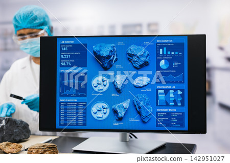 Laboratory worker in protective gear studies mineral samples beside a computer screen blue data charts and rock analysis results. Laboratory worker in protective gear studies mineral samples beside a computer screen blue data charts and rock analysis results. 142951027